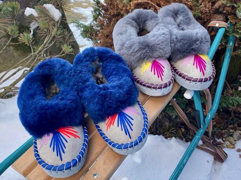 May include: Two pairs of cozy, handcrafted slippers. One pair features vibrant blue fur trim, while the other has gray fur. Both have embroidered designs in red, blue, and yellow on a light gray base, with stitched edges.