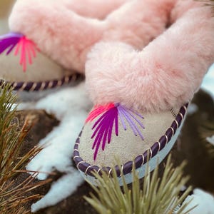 May include: A pair of light-colored slippers with pink faux fur trim. The slippers have a decorative stitched design in shades of pink and purple. The edges are trimmed with a dark purple cord. The slippers are resting on a rock with snow and pine needles in the background.