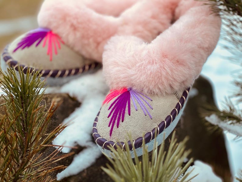 May include: A pair of light-colored slippers with pink faux fur trim. The slippers have a decorative stitched design in shades of pink and purple. The edges are trimmed with a dark purple cord. The slippers are resting on a rock with snow and pine needles in the background.