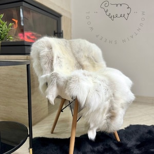 May include: A white sheepskin rug with brown accents is draped over a wooden chair. The rug is sitting on a black sheepskin rug. The text "Mountain Sheep Wool" is written in a circle around a sheep illustration.
