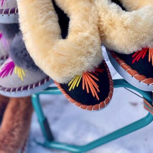 May include: Close-up of several pairs of colorful, embroidered slippers. The slippers have a black base with orange, yellow, and red stitching. They feature a light brown, fluffy trim and a leather-like sole. The slippers are displayed on a teal stand.
