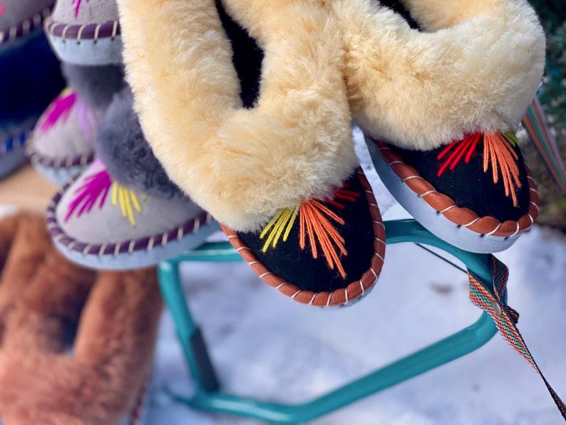 May include: Close-up of several pairs of colorful, embroidered slippers. The slippers have a black base with orange, yellow, and red stitching. They feature a light brown, fluffy trim and a leather-like sole. The slippers are displayed on a teal stand.