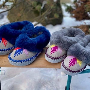 May include: Two pairs of cozy, sheepskin slippers. One pair is navy blue with colorful embroidery, the other is gray. Both have a light gray suede base and are sitting on a wooden surface. The slippers are designed for indoor use.