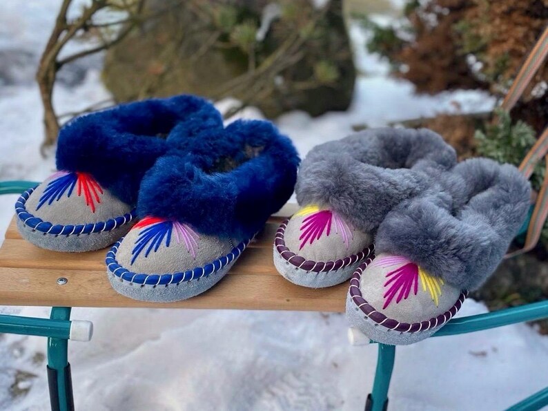 May include: Two pairs of cozy, sheepskin slippers. One pair is navy blue with colorful embroidery, the other is gray. Both have a light gray suede base and are sitting on a wooden surface. The slippers are designed for indoor use.