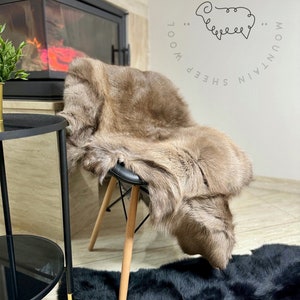 May include: A brown sheepskin rug with long, soft fur is draped over a black chair with wooden legs. The rug is sitting on a black faux fur rug. The text "MOUNTAIN SHEEP WOOL" and a stylized sheep are visible in the background.