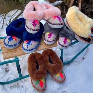 May include: A collection of colorful, embroidered sheepskin slippers in various colors, including blue, pink, gray, and brown. The slippers are displayed on a green sled in a snowy outdoor setting. Each slipper features a unique embroidered design.