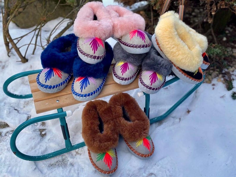 May include: A collection of colorful, embroidered sheepskin slippers in various colors, including blue, pink, gray, and brown. The slippers are displayed on a green sled in a snowy outdoor setting. Each slipper features a unique embroidered design.