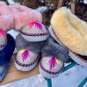 May include: A collection of colorful, embroidered sheepskin slippers. The slippers feature a light-colored suede base with decorative stitching and a vibrant, stylized floral design. The fluffy tops come in pink, gray, blue, and yellow.