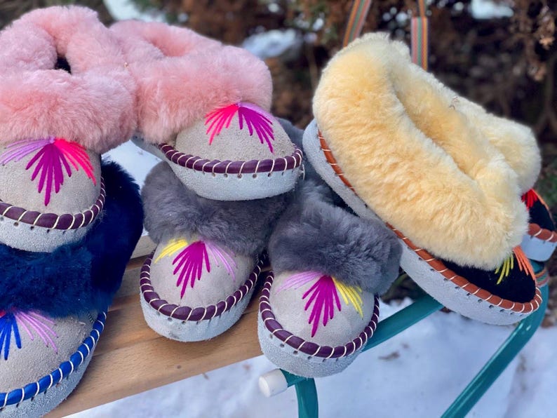 May include: A collection of colorful, embroidered sheepskin slippers. The slippers feature a light-colored suede base with decorative stitching and a vibrant, stylized floral design. The fluffy tops come in pink, gray, blue, and yellow.