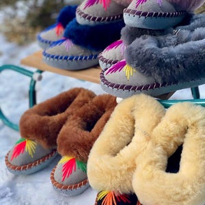 May include: A collection of colorful, embroidered sheepskin slippers. The slippers come in various colors, including pink, blue, gray, brown, and yellow. Each slipper features a suede base with decorative stitching and a vibrant, embroidered design.