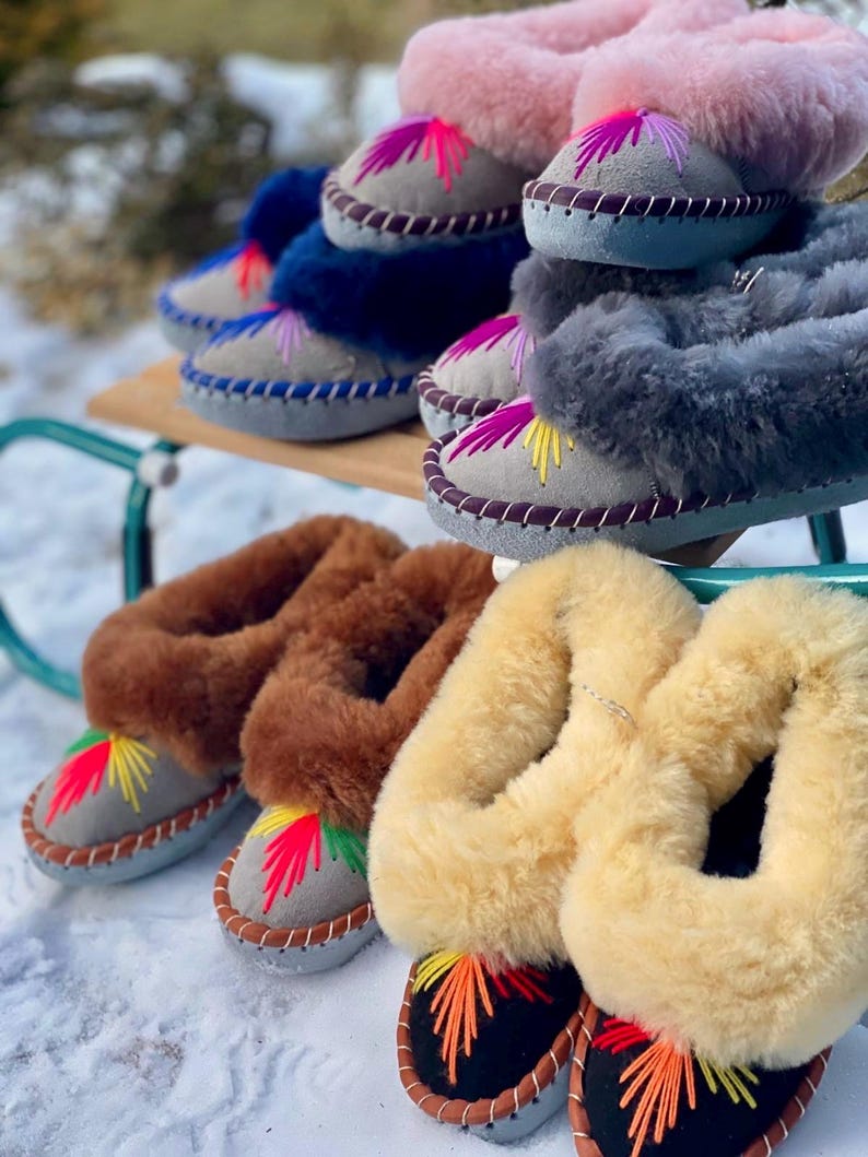 May include: A collection of colorful, embroidered sheepskin slippers. The slippers come in various colors, including pink, blue, gray, brown, and yellow. Each slipper features a suede base with decorative stitching and a vibrant, embroidered design.