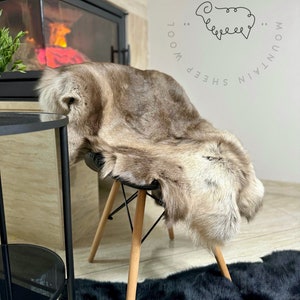 May include: A brown and white sheepskin rug with a black sheep silhouette logo that says "Mountain Sheep Wool Derisi" on a white background. The rug is draped over a wooden chair with black legs. The chair is sitting on a black faux fur rug.
