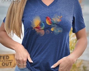 Puede incluir: Una camiseta azul con un diseño de acuarela de dos pájaros coloridos posados en una rama. El diseño está en tonos de amarillo, naranja, rojo, azul y verde.