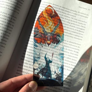 Op de afbeelding: Een bladwijzer met een kleurrijk glas-in-lood ontwerp met een rode demon en een blauwe figuur die een staf vasthoudt. De bladwijzer heeft de vorm van een puntige boog.