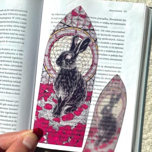 Può includere: Un'illustrazione in bianco e nero di un coniglio seduto su un letto di fiori rosa e bianchi. Il coniglio è circondato da un disegno circolare con un motivo geometrico. L'illustrazione è stampata su un segnalibro.