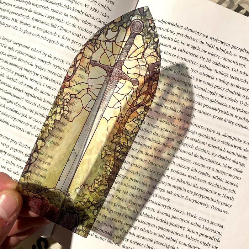 May include: A colorful glass bookmark featuring a sword and a tree with green and brown leaves. The bookmark is shaped like a sword and has a stained glass look.