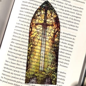 May include: A stained glass bookmark featuring a sword with a golden hilt and a cross guard. The sword is surrounded by a colorful stained glass design of trees and leaves. The bookmark is perfect for book lovers and fantasy fans.