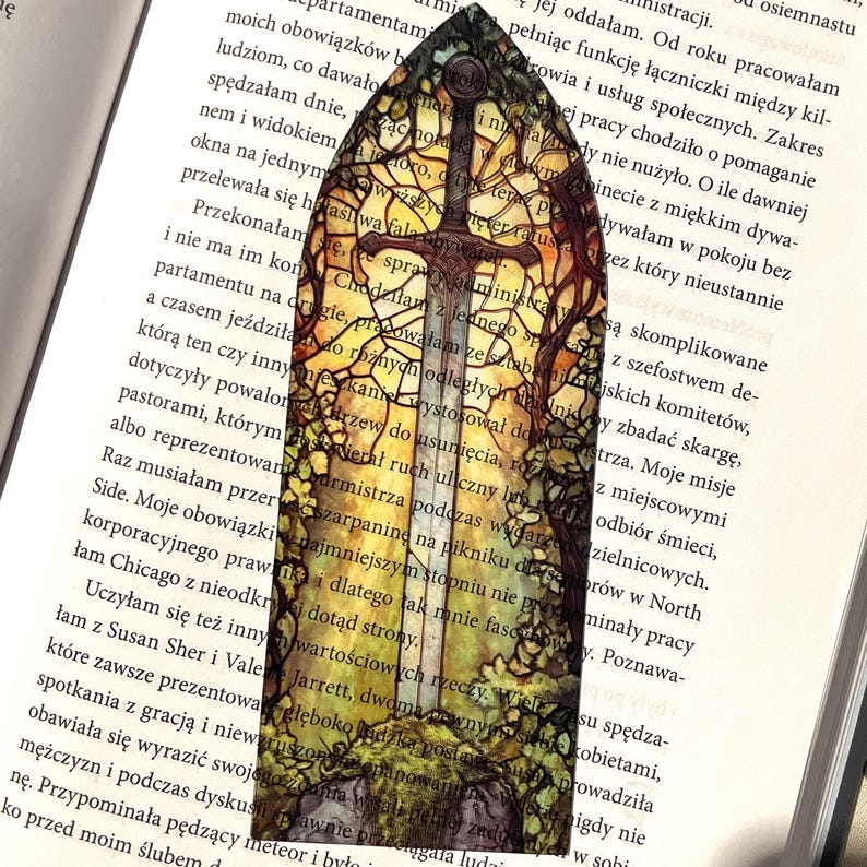 May include: A stained glass bookmark featuring a sword with a golden hilt and a cross guard. The sword is surrounded by a colorful stained glass design of trees and leaves. The bookmark is perfect for book lovers and fantasy fans.