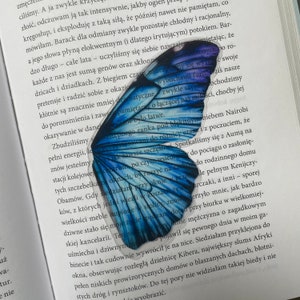 Può includere: Un segnalibro a forma di farfalla blu con un bordo bianco. Il segnalibro è su una pagina di un libro con testo su un viaggio in Kenya.