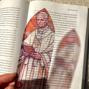 May include: A stained glass bookmark depicting a man in a white robe with a gold cross. He is holding a book in his hands. The bookmark is shaped like a pointed arch.