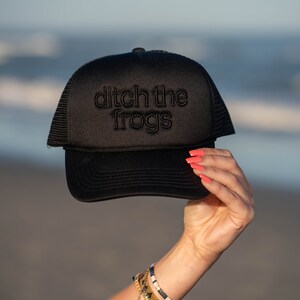 Puede incluir: Una gorra de camionero negra con una parte trasera de malla negra. La gorra tiene el texto "ditch the frogs" bordado en blanco en la parte delantera.