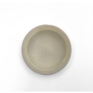 May include: A circular, light beige, shallow dish with a smooth, matte finish. The object features a wide, flat rim and a concave center with a single, small, centered hole at the base, presenting a simple, functional, and unadorned design.