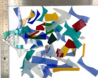 May include: A collection of assorted stained glass pieces in various shapes and colors, including blue, yellow, red, green, and white, arranged next to a metal ruler showing measurements up to 12 inches and a small circular coin for scale.