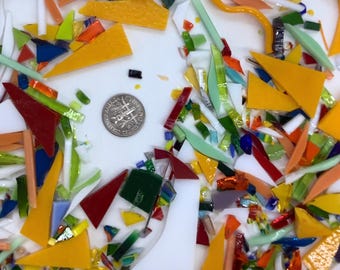 May include: A collection of multicolored glass shards in various shapes and sizes, including yellow, white, green, blue, orange, and red pieces, scattered around a silver United States dime coin placed in the center for scale reference.