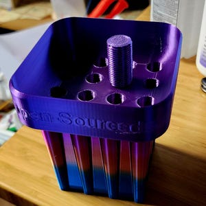May include: A 3D-printed pen holder with a gradient of blue, orange, and purple. The top is a solid purple with several circular holes and a central, threaded post. The text "Open Source" is visible on the side.