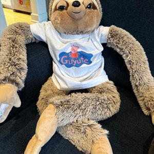 Puede incluir: Un juguete de peluche de perezoso marrón y beige que lleva una camiseta blanca con un logotipo azul y rosa que dice "Guyute".