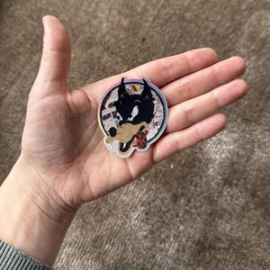 May include: A round sticker featuring a black and white wolf illustration. The wolf is surrounded by a colourful, abstract design. The sticker is on a hand.