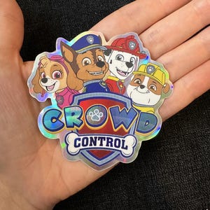 May include: A holographic sticker featuring the Paw Patrol characters Chase, Skye, Marshall, and Rubble. The sticker has a blue, red, yellow, and pink color scheme. The text "Crowd Control" is printed on the sticker.