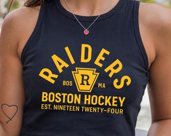 May include: Black crop top with the text "RAIDERS BOSTON HOCKEY EST. NINETEEN TWENTY-FOUR" in yellow. The top has a racerback design and a round neckline. A small heart pendant necklace is visible.