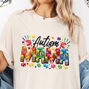Könnte beinhalten: Cremefarbenes T-Shirt mit den Worten "Autism MAMA" in bunten Buchstaben und Puzzleteil-Designs. Das Shirt hat auch Handabdrücke und Blumenakzente. Der Text "Mother's Day Gift" wird auf einem roten Rechteck angezeigt.