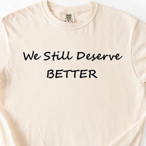 May include: A cream-coloured t-shirt featuring the black text "We Still Deserve BETTER". The text is centred. The shirt has a crew neck and short sleeves. The fabric appears soft and slightly creased.
