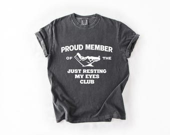 May include: Dark gray t-shirt with rolled-up sleeves. The shirt features white text that reads "PROUD MEMBER OF THE JUST RESTING MY EYES CLUB" with a graphic of a person reclining in a chair.