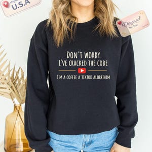 Könnte beinhalten: Schwarzes Sweatshirt mit Rundhalsausschnitt und dem weißen Text "DON'T WORRY I'VE CRACKED THE CODE" und "I'M A COFFEE A TIKTOK ALORRTHIM". Das Design enthält eine YouTube-Play-Button-Grafik. Das Sweatshirt wird mit blauen Jeansshorts kombiniert.