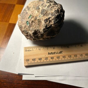 Könnte beinhalten: Ein rauer, grauer und brauner Stein mit grünen Akzenten. Der Stein hat einen Durchmesser von etwa 5 cm und liegt auf einer weißen Oberfläche.
