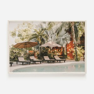 May include: A tropical resort pool scene with lounge chairs under palm trees and umbrellas. The pool is surrounded by lush greenery and a red building in the background.