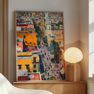 Puede incluir: Una impresión enmarcada de una pintura colorida que representa una escena de calle de ciudad con edificios, árboles y personas. La pintura está realizada en un estilo estilizado, casi de dibujos animados, con colores brillantes y líneas audaces.