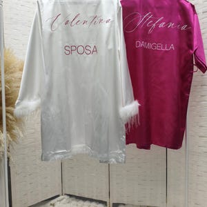 Può includere: Una vestaglia in raso bianco con polsini in piuma che riporta il testo Valentina e SPOSA in rosso scuro, accanto a una vestaglia in raso magenta con il testo Stefania e DAMIGELLA in bianco, entrambe appese a un appendiabiti bianco.