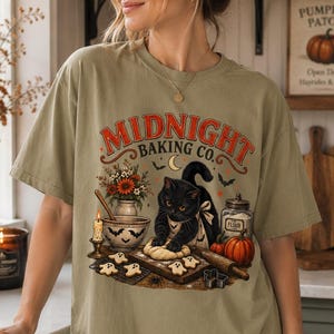 May include: A sage green short-sleeve t-shirt featuring a graphic print of a black cat wearing an apron and kneading dough. The text reads Midnight Baking Co. and includes illustrations of pumpkins, a candle, a flour jar, and ghost-shaped cookies.