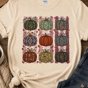 May include: A cream-coloured short-sleeve t-shirt featuring a graphic print of nine pumpkins arranged in a grid. Each pumpkin displays a unique pattern, including floral, geometric, and mushroom motifs in various autumn-inspired colours.