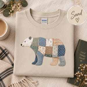 Puede incluir: Una sudadera de cuello redondo color arena con un gráfico central de un oso compuesto por varios patrones de tela de patchwork en tonos azul, verde, beige y blanco. La etiqueta de la marca Gildan es visible en el cuello interior.