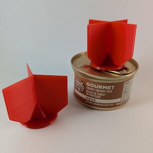 Op de afbeelding: Een rode, 3D-geprinte deksel voor kattenvoerblikjes en een blik Gourmet Chunky Chicken Feast paté. De deksel is ontworpen om op de bovenkant van het blik te passen. Het blik heeft tekst in het Engels en Frans.