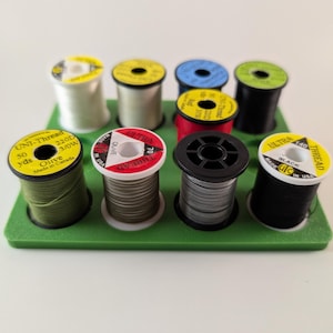 Puede incluir: Una colección de carretes de hilo en varios colores, incluyendo verde oliva, negro y blanco, dispuestos en una bandeja verde. Los carretes tienen etiquetas amarillas con texto negro, y algunos tienen detalles en rojo o azul. El hilo está etiquetado como "UNI-Thread" y "ULTRA Thread".