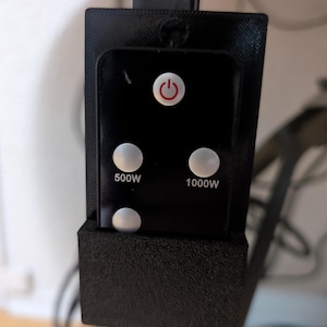 Puede incluir: Un panel de control negro con un botón de encendido rojo y tres botones blancos. El panel muestra el texto "500W" y "1000W". El panel está montado en un soporte negro.