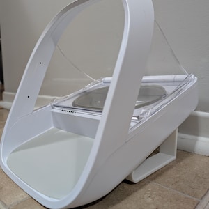 Puede incluir: Dispensador de comida para mascotas blanco con una cubierta transparente en forma de arco. El dispensador tiene una base gris y un cuenco de acero inoxidable. El diseño es moderno e inclinado, con una pata de soporte.