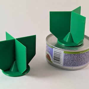 Op de afbeelding: Twee groene plastic dekselopeners voor blikken. De grotere is op een blikje kattenvoer. De kleinere is op een wit oppervlak.