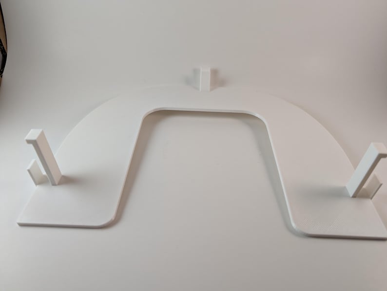 May include: A white, U-shaped object with a flat base and curved top. Two rectangular supports extend from the base, and a third support rises from the center of the top. The object appears to be made of a solid, smooth material.
